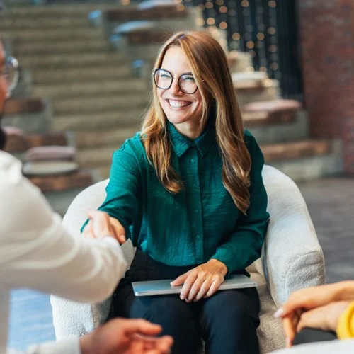 VMS Workforce Solutions image Business person sat in an armchair, holding a laptop in one hand and shaking a colleagues hand with the other