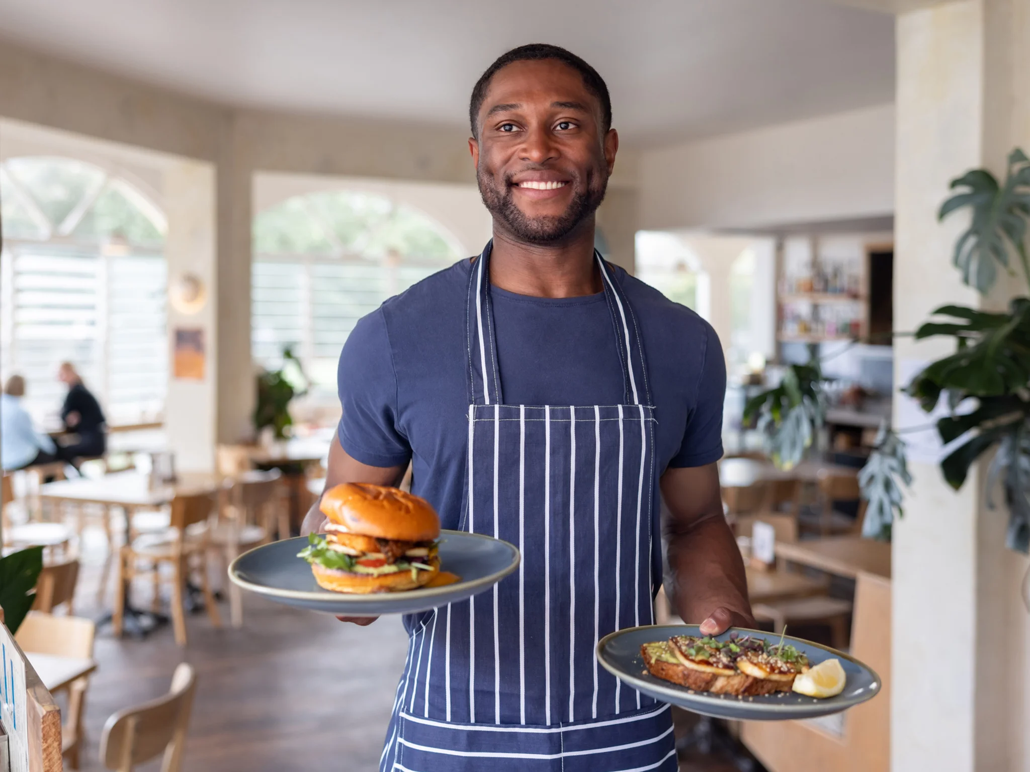 A smiling waiter wearing an apron carrying two plates of food