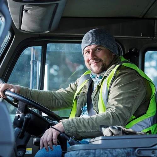HGV Driver Training image HGV driver sat behind the wheel in a yellow hi vis jacket and wearing a beanie hat