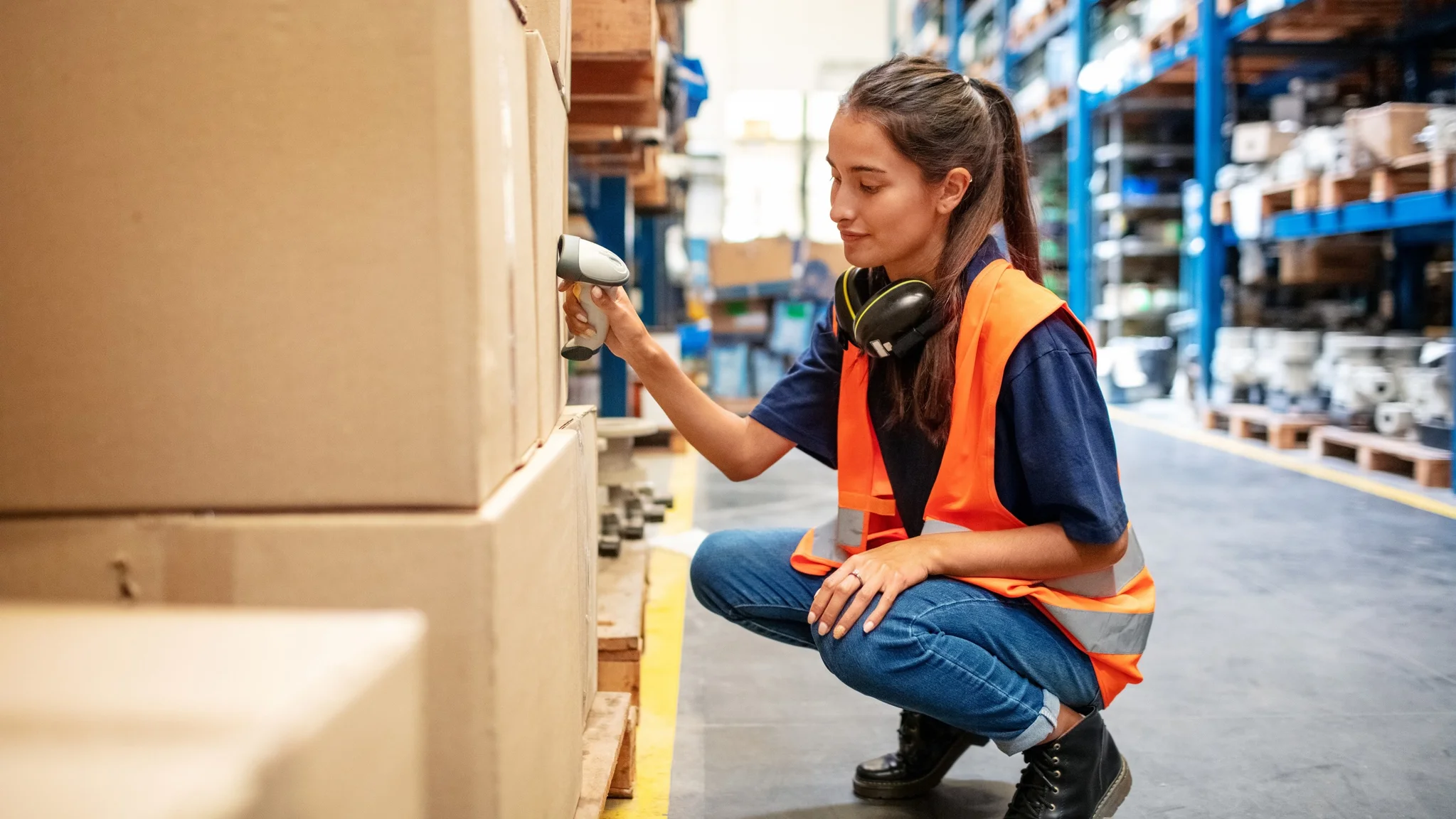Warehouse Worker wearing an orange hi vis vest. They have ear defenders around their neck and are in a warehouse crouching down to scan a cardboard box.