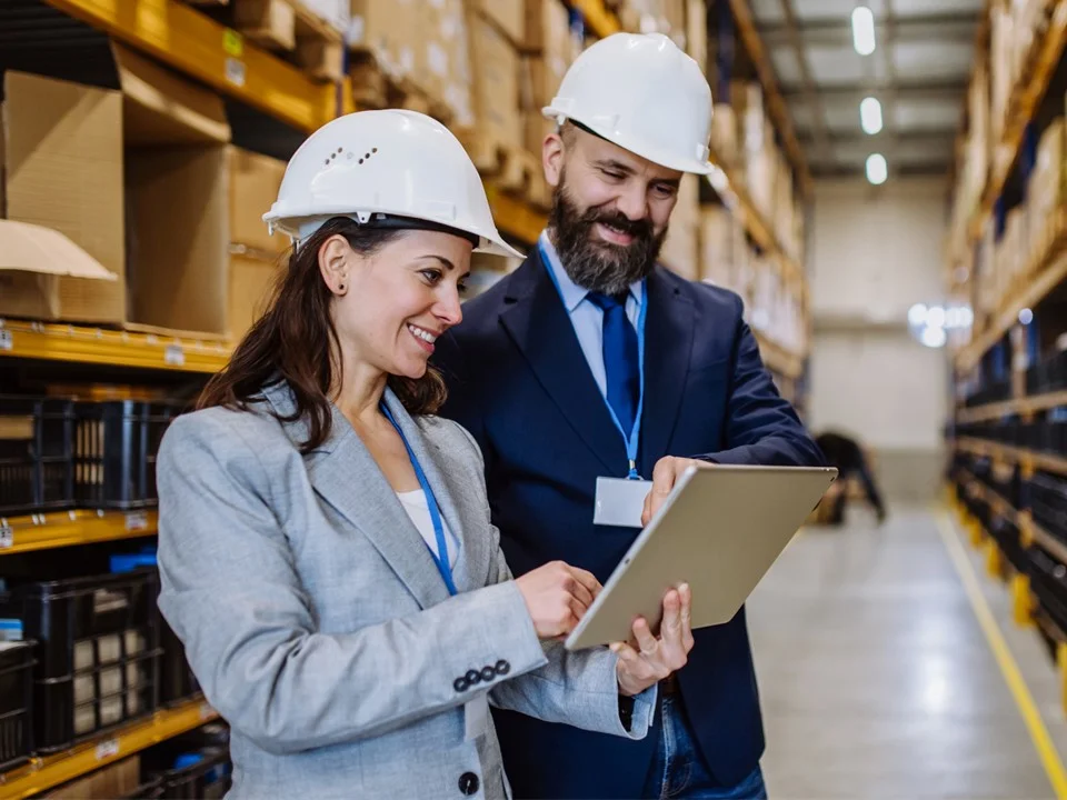Two warehouse workers wearing suits and a hardhat, looking at a tablet.