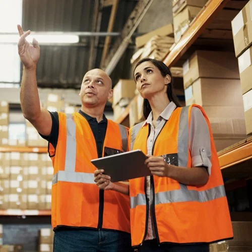 Two warehouse workers wearing orange hi vis jackets, stood in a warehouse surrounded by boxes