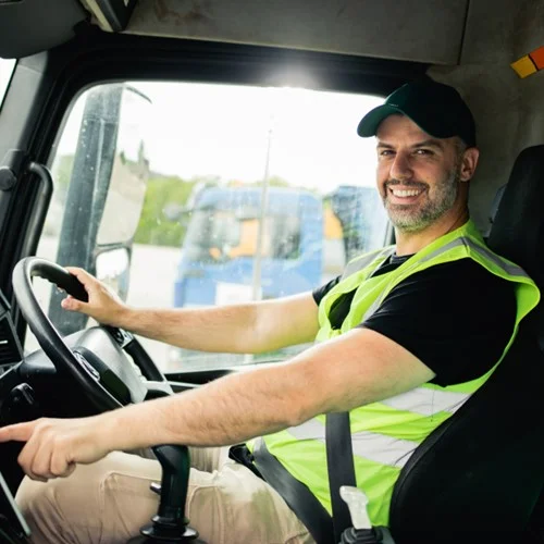 Permanent Recruitment image A Driver in a yellow hi vis vest and wearing a cap sat in an HGV with one hand on the steering wheel
