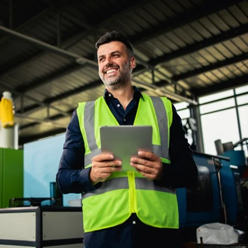 RecruiterBot image A Warehouse Manager in a yellow hi vis vest, stood in a warehouse whilst holding a tablet