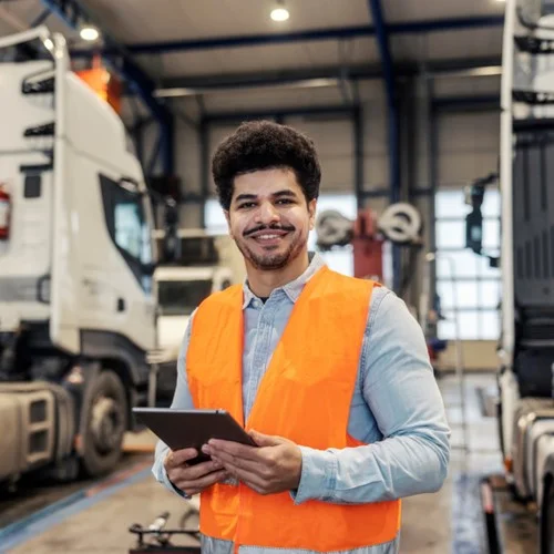 A Logistics Manager stood in an HGV depot wearing an orange hi vis jacket and holding a tablet