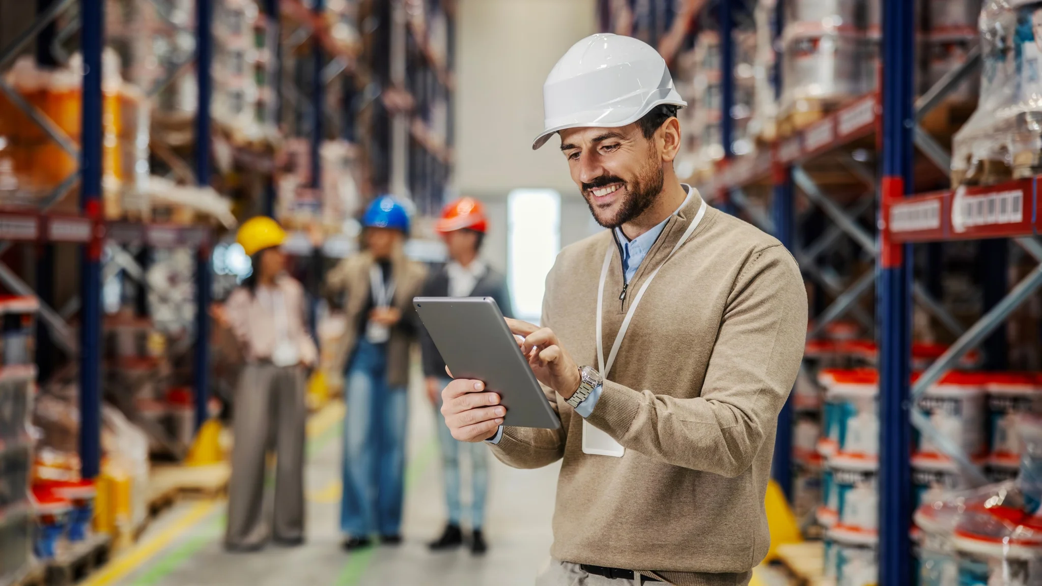 A Supply Chain Planner stood in a warehouse. They are wearing a white hard hat, a lanyard, and they are looking at a tablet.
