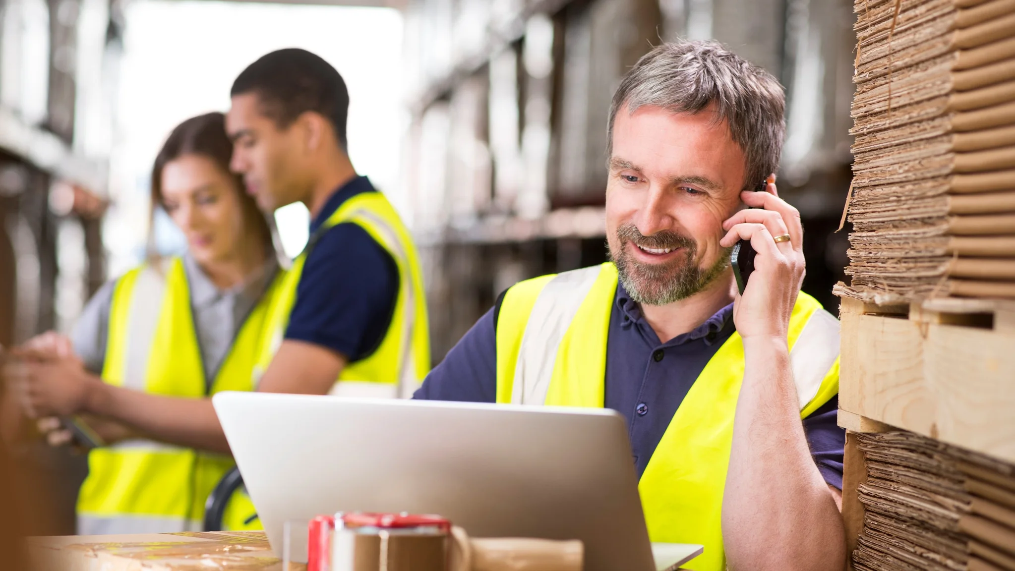 Logistics Coordinator in hi vis vest sat at a laptop whilst talking on a mobile phone