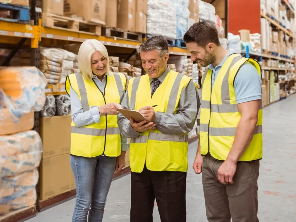 Three warehouse workers in yellow hi-vis vests stood in a warehouse looking at a clipboard