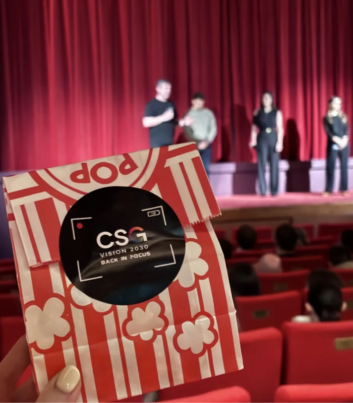 bag of branded CSG Talent popcorn with blurred background. four people are in the background on a theatre stage