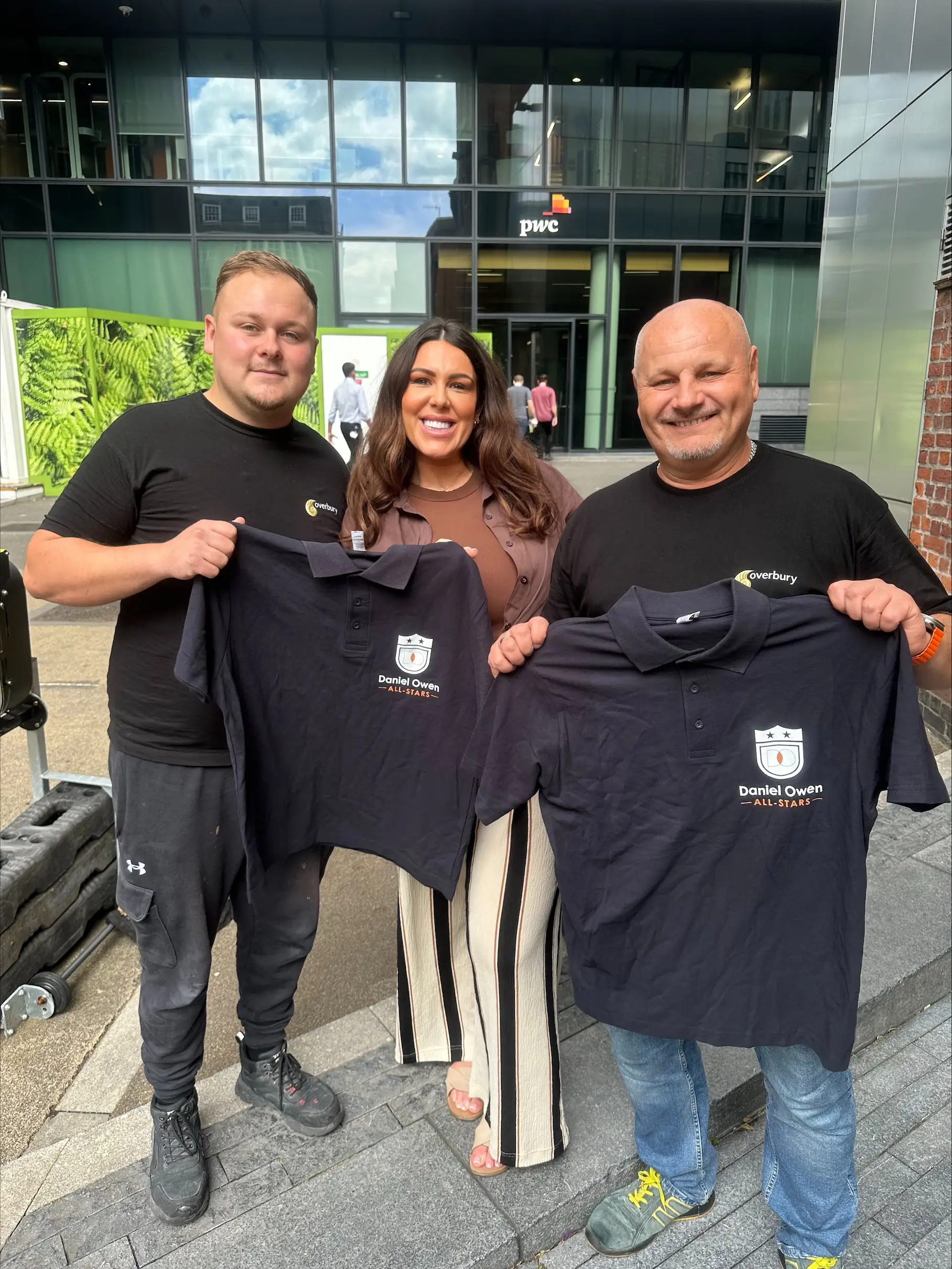 Three individuals stood in front of an office building. Two male, one female. The female individual is rewarding the two construction site workers with some branded Daniel Owen merchandise for their hard work on construction sites.