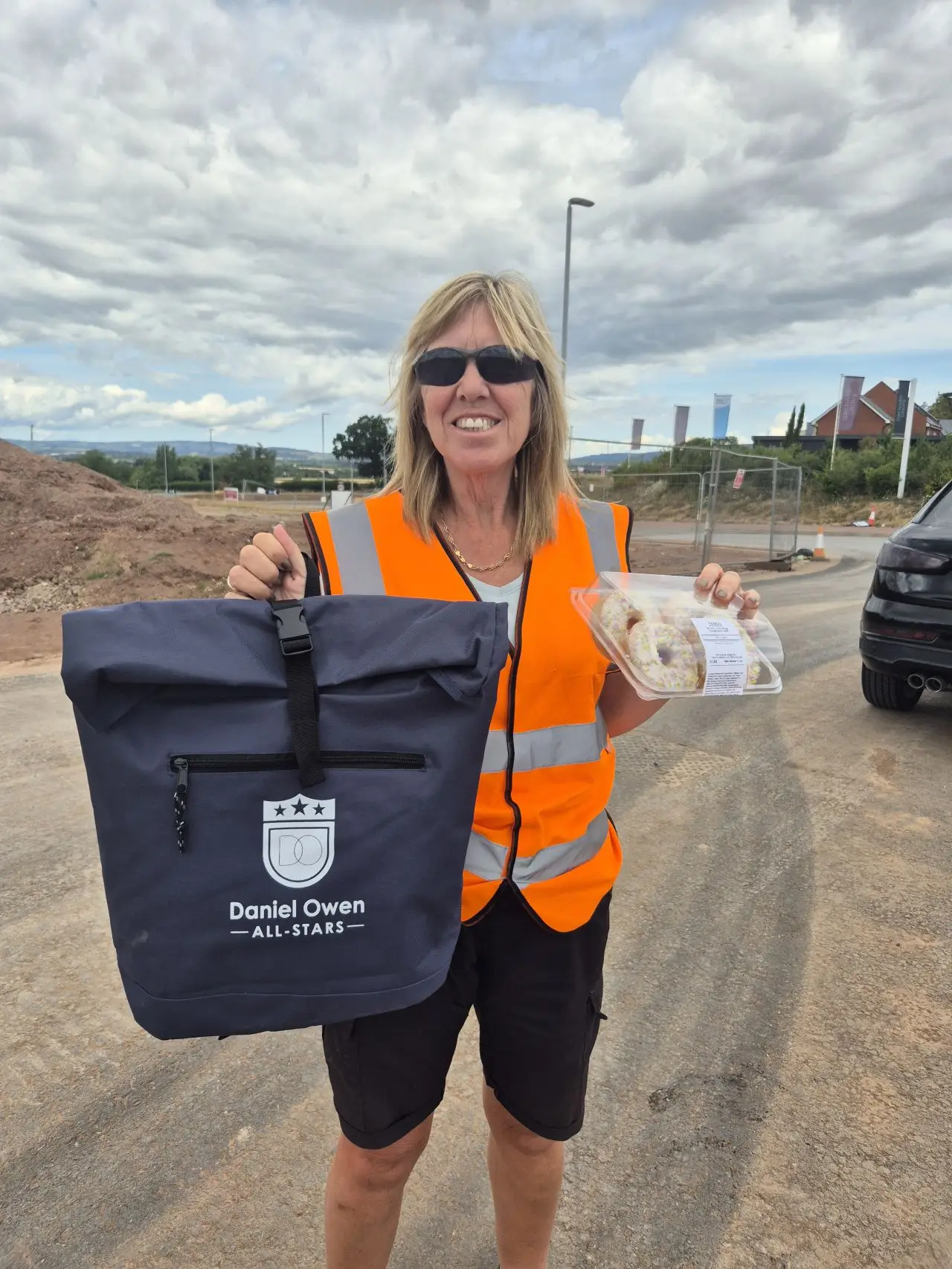 A female construction worker in an orange hi-vis holding their rewards for hard work, a bag and a box of donuts.