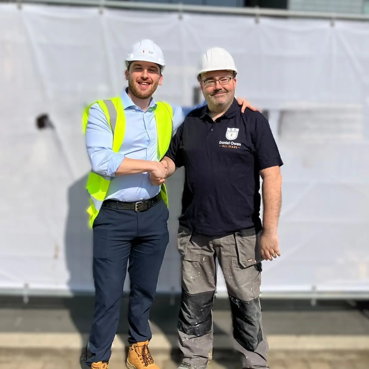 Two males standing together together, both with white hard-hats on. One is in a suit and a hi-vis, the other is a construction clothing. They are shaking hands as the male in the suit is rewarding the construction worker for his hard work. 
