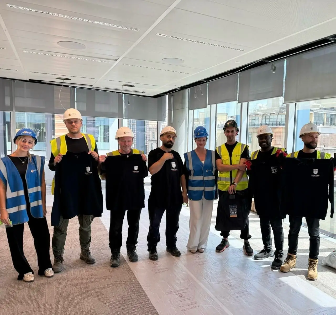 A large group of construction workers, a mix of male and female all stood together holding up their Daniel Owen branded t-shirts rewarded to them as part of a candidate recognition scheme for hard work on site.