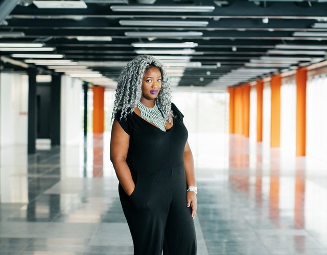 A photo of Dr Anne-Marie Imafidon, MBE, a successful business woman and founder of a social enterprise that supports women in STEM careers. She is stood in an office space wearing a black jumpsuit and an elaborate silver necklace. She has curly grey hair and is wearing purple lipstick.