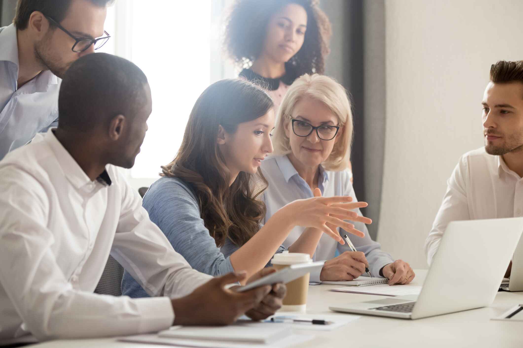 An image of a group of colleagues sitting around a desk looking at a laptop. There is a woman sat in the middle of the group, and she looks to be leading the conversation while looking at a laptop. Her colleagues are looking at her, as if listening intently.