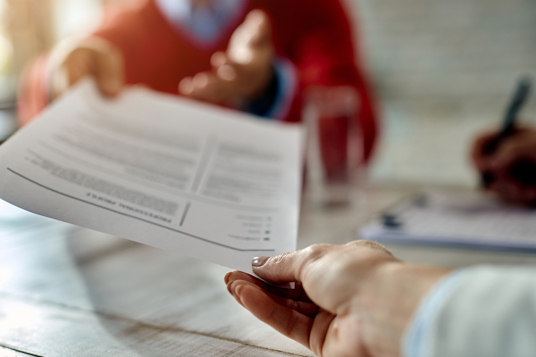 An image of two people. One person is handing a CV to the other person across a table. You can only see their hands, the focus is on the CV.