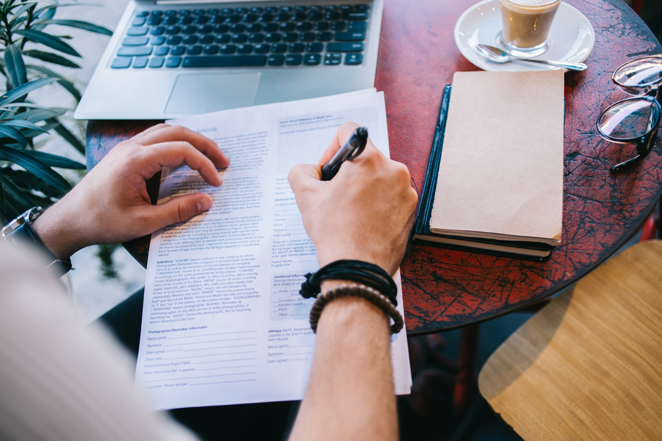 A person is sat at a table at a coffee shop. You can see a laptop and notepads on their table, along with a coffee. They are filling in a form, possibly an application form.