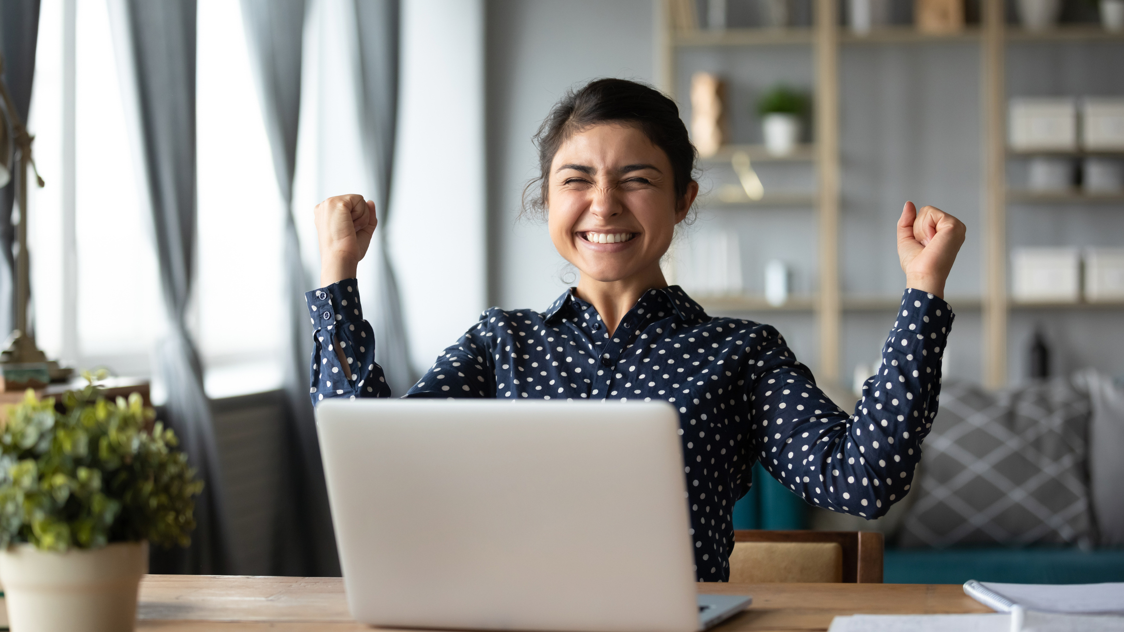 A photo of a woman sitting at a desk with a laptop in front of her. She is smiling and raising her hands in the air as if celebrating.