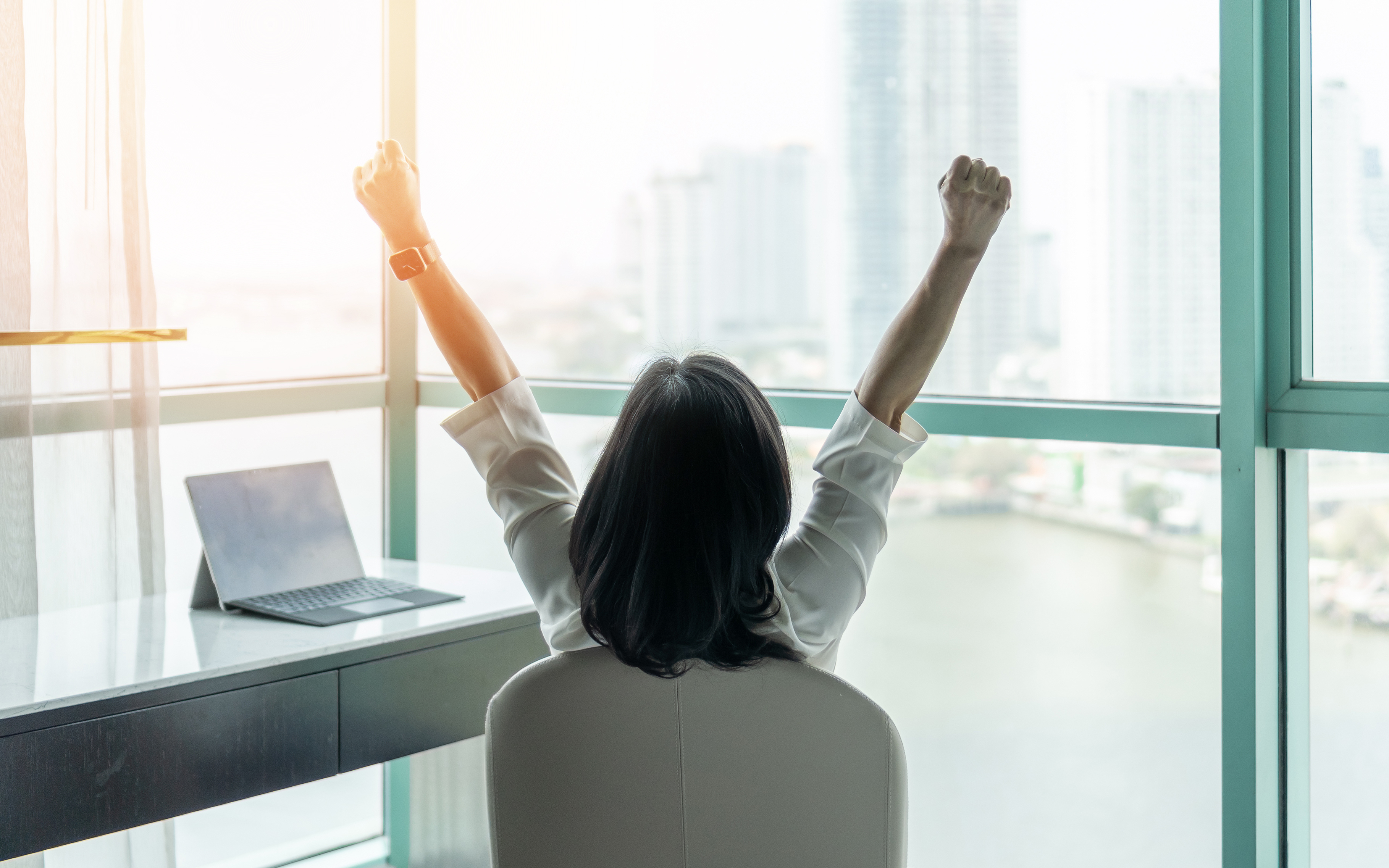 A photo of a woman sitting at a desk. She is sat on a chair and her back is facing the camera. Her arms are stretching in the air as if stretching after a long day of work.