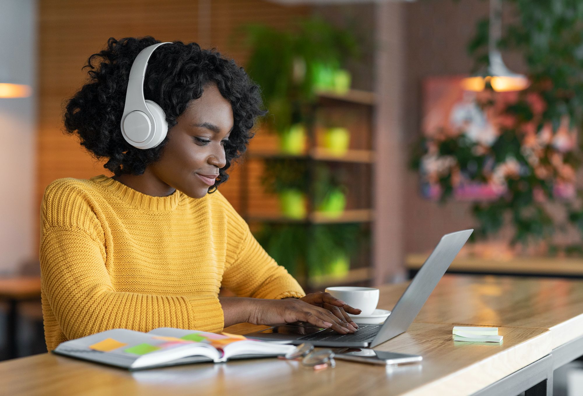 A photo of a woman working on a laptop at a desk. She has headphones on and is smiling at her laptop.