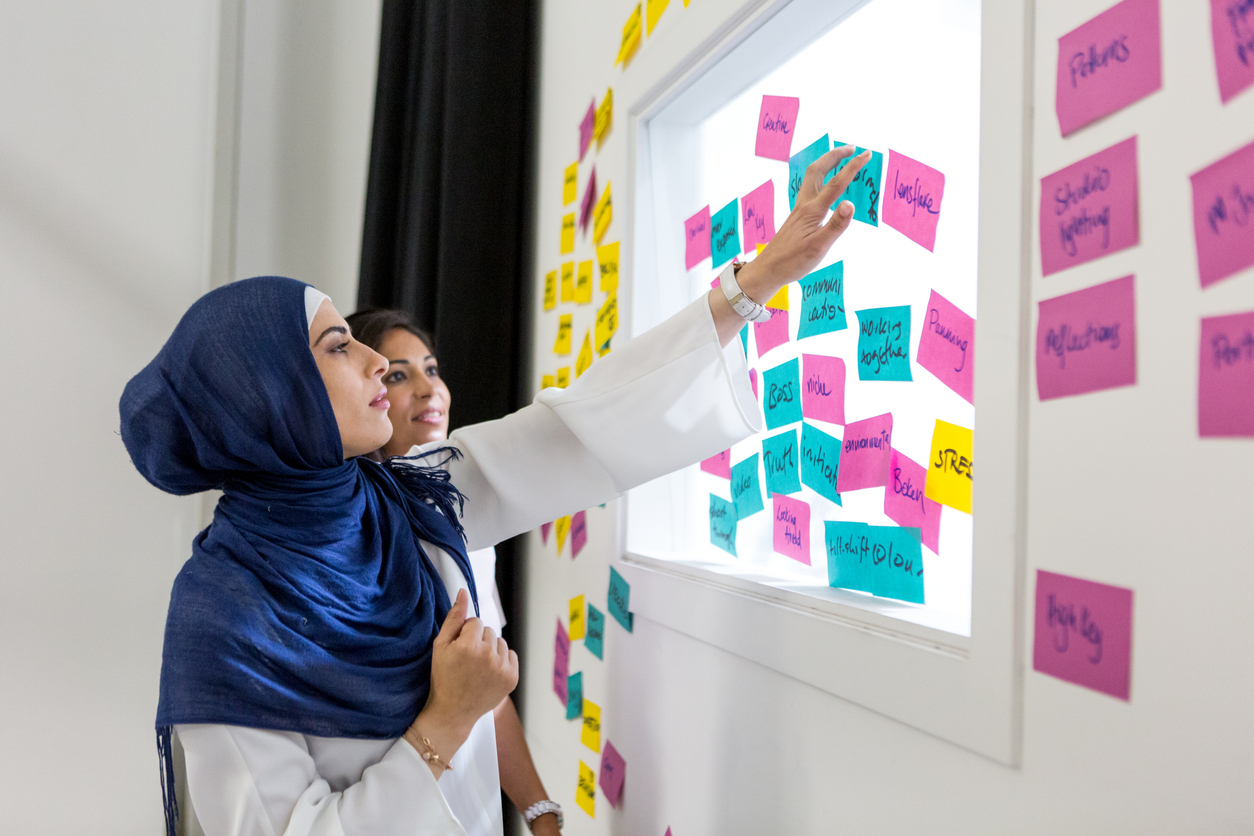 Two female colleagues in the workplace. Both women are brown, and one is wearing a hijab. They are both looking at a display which has post-it notes on.