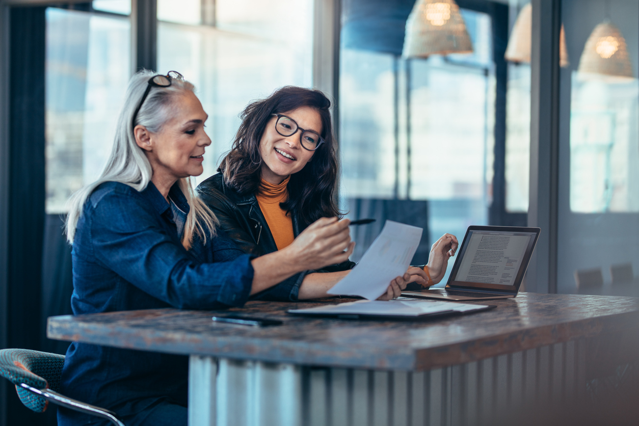 Two professional women sat side by side at a desk. There is a laptop on the desk, and one woman is holding a piece of paper. Both women are looking at the piece of paper smiling.