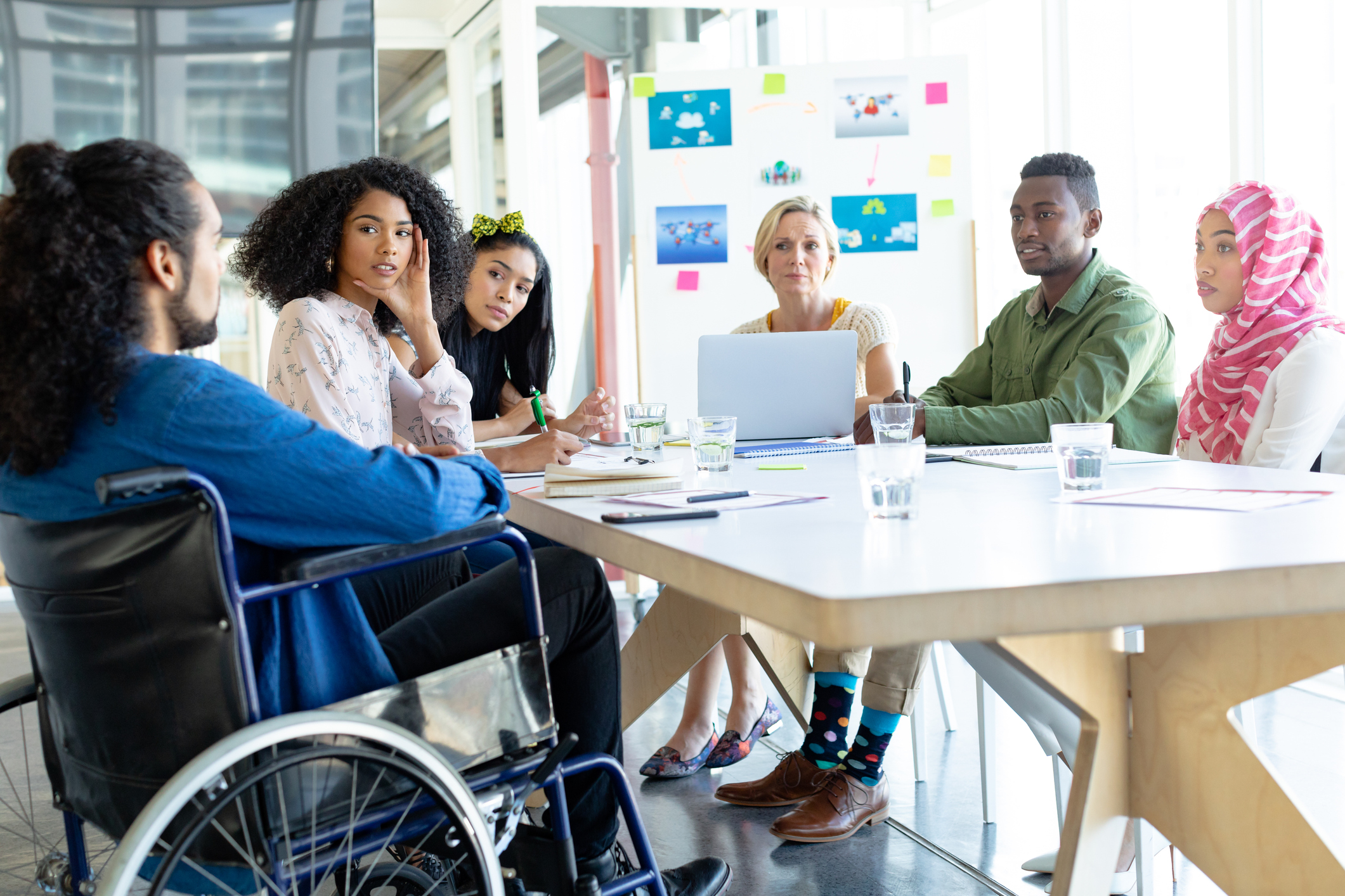 A group of diverse people are sat around a table. There are men and women, a man in a wheelchair, a woman in a hijab, and people of different races, Everyone is looking at the man in a wheelchair who appears to be talking to the group. Some people ate taking notes on a notepad and on a laptop.