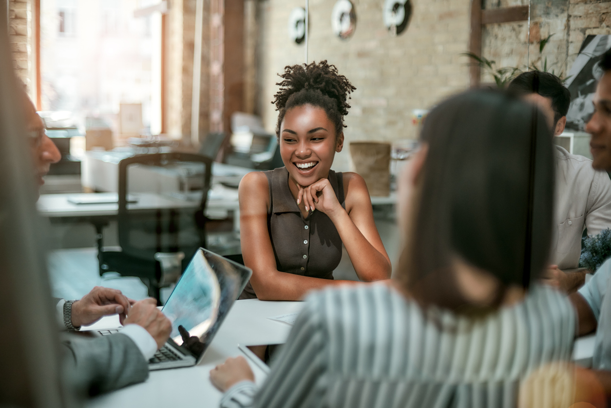 A woman is sat opposite her colleagues at a table. They appear to be in a work environment. The woman is laughing and smiling, and appears to be happy in her place of work.