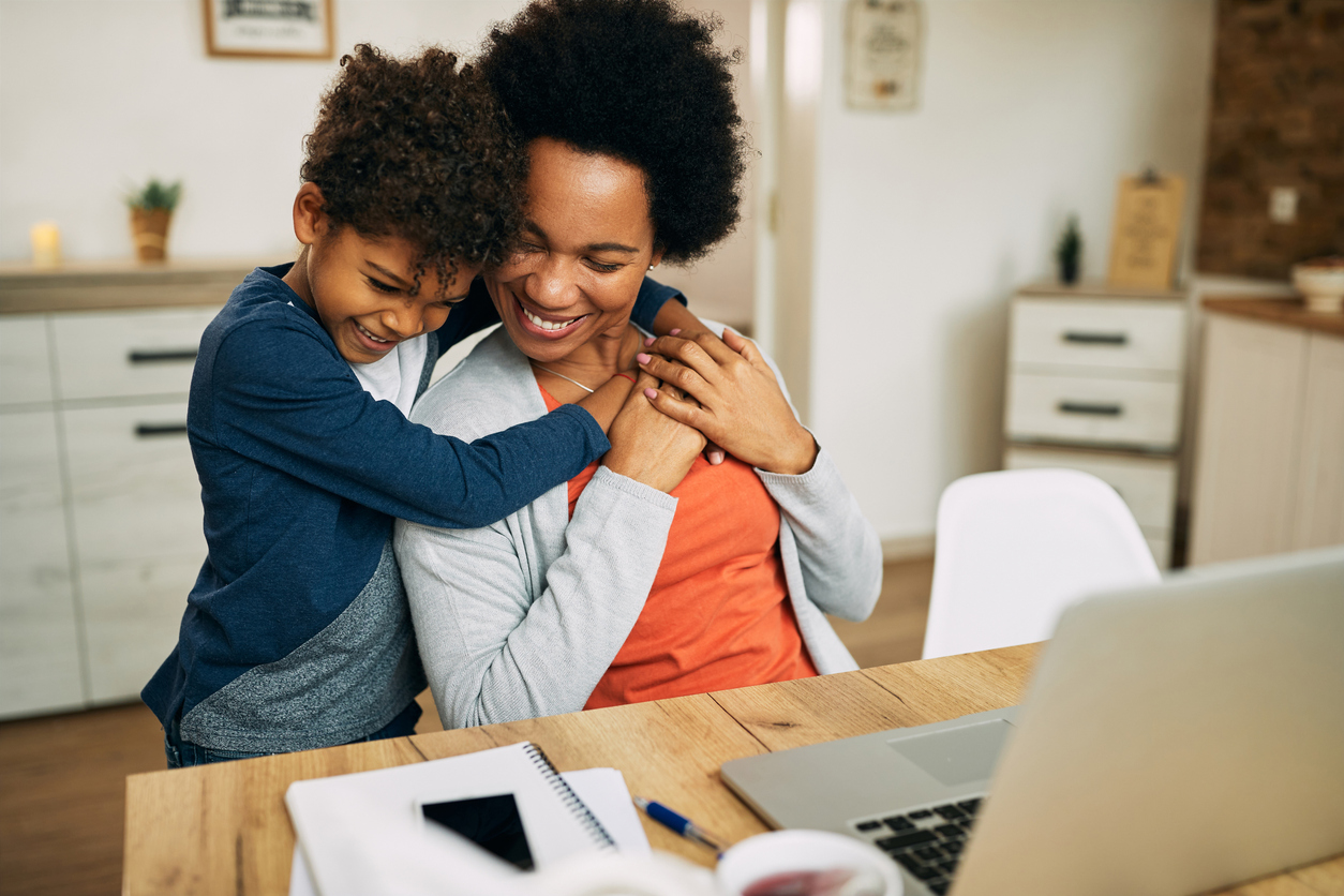 A woman working from home. She is sat at a table with a laptop and notepad in front of her, A young boy is hugging her while she is sat at the desk. They are both smiling.