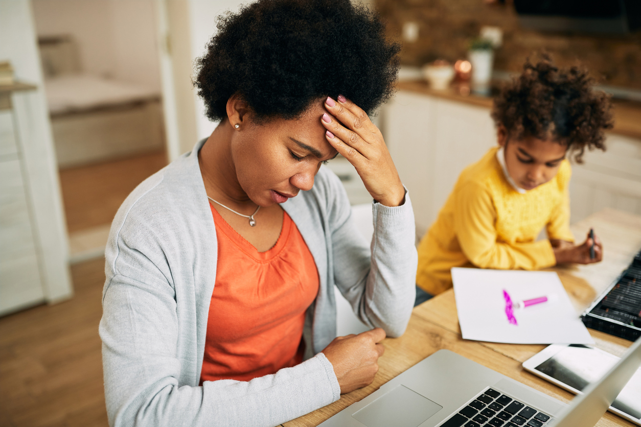 A woman is sat at a desk with a laptop in front of her. She has her head in her hands and looks stressed. A young boy is sat beside her doing some colouring, suggesting the woman is working from home.