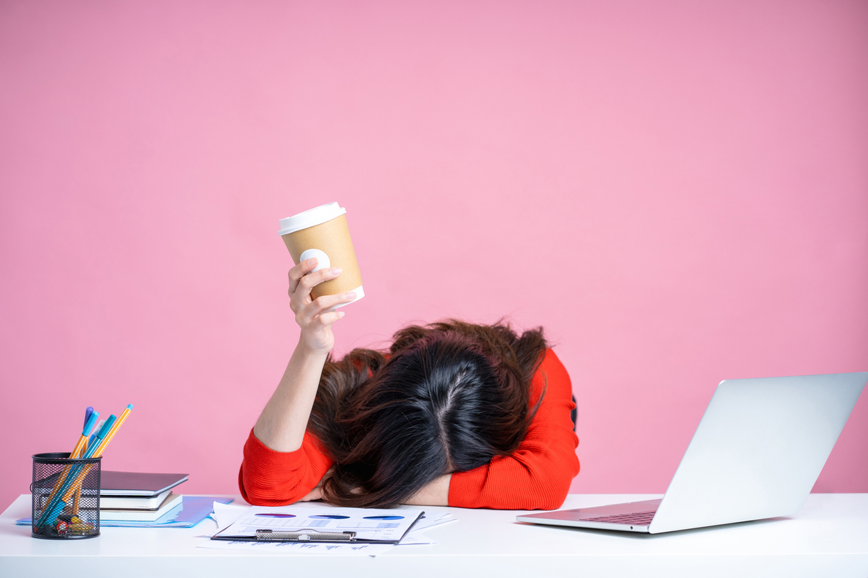 A woman is sat at a desk. There is a laptop on the desk, along with a pen pot with pens inside, a pile of books and a clipbpard with paper on. The woman has her face in her arm on the table, as if fed up or tired. She is also holding a coffee cup.