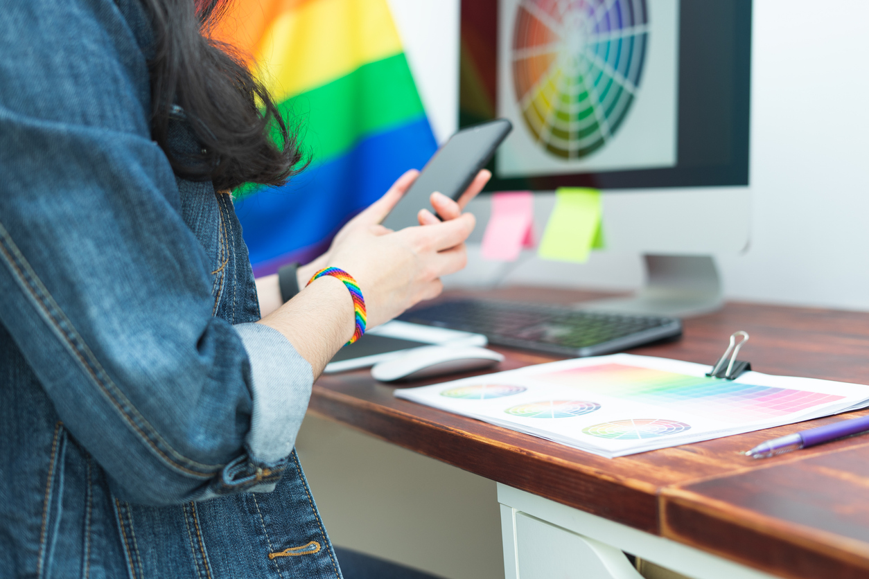 A woman wearing a denim jacket and a multicoloured pride wristband is stood at a desk looking at a mobile phone.