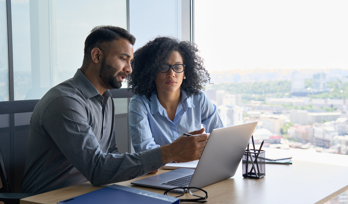 A man and a woman are sat at a desk, looking at a laptop. They are dressed smartly and in an office setting. The man is pointing to the laptop, as if explaining something to the woman.