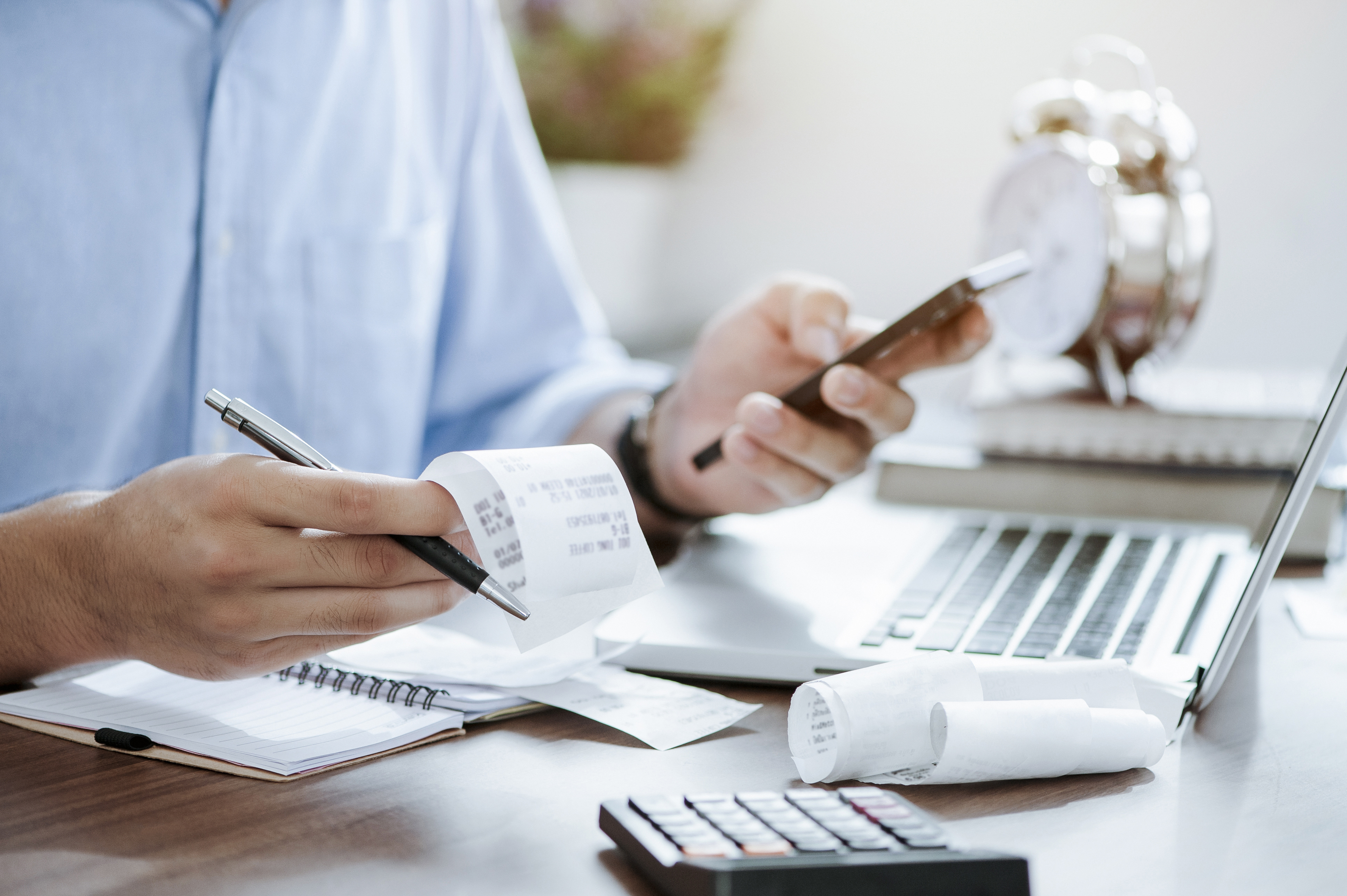 A professional person dressed in a shirt is sat at a desk. You can not see their face but can see their hands. They have a pen and a receipt in one hand and a phone in the other. There is a calculator and laptop on the desk. It looks as if they are calculating some expenses.