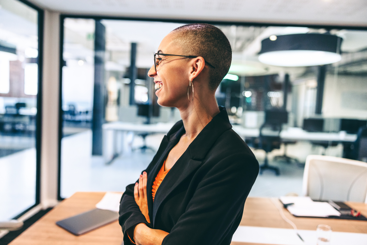 A woman is stood in an office environment, and dressed in smart suit wear. She has glasses, a shaved head and is wearing hoop earrings. She is looking off camera to the side, and appears to be smiling at someone.