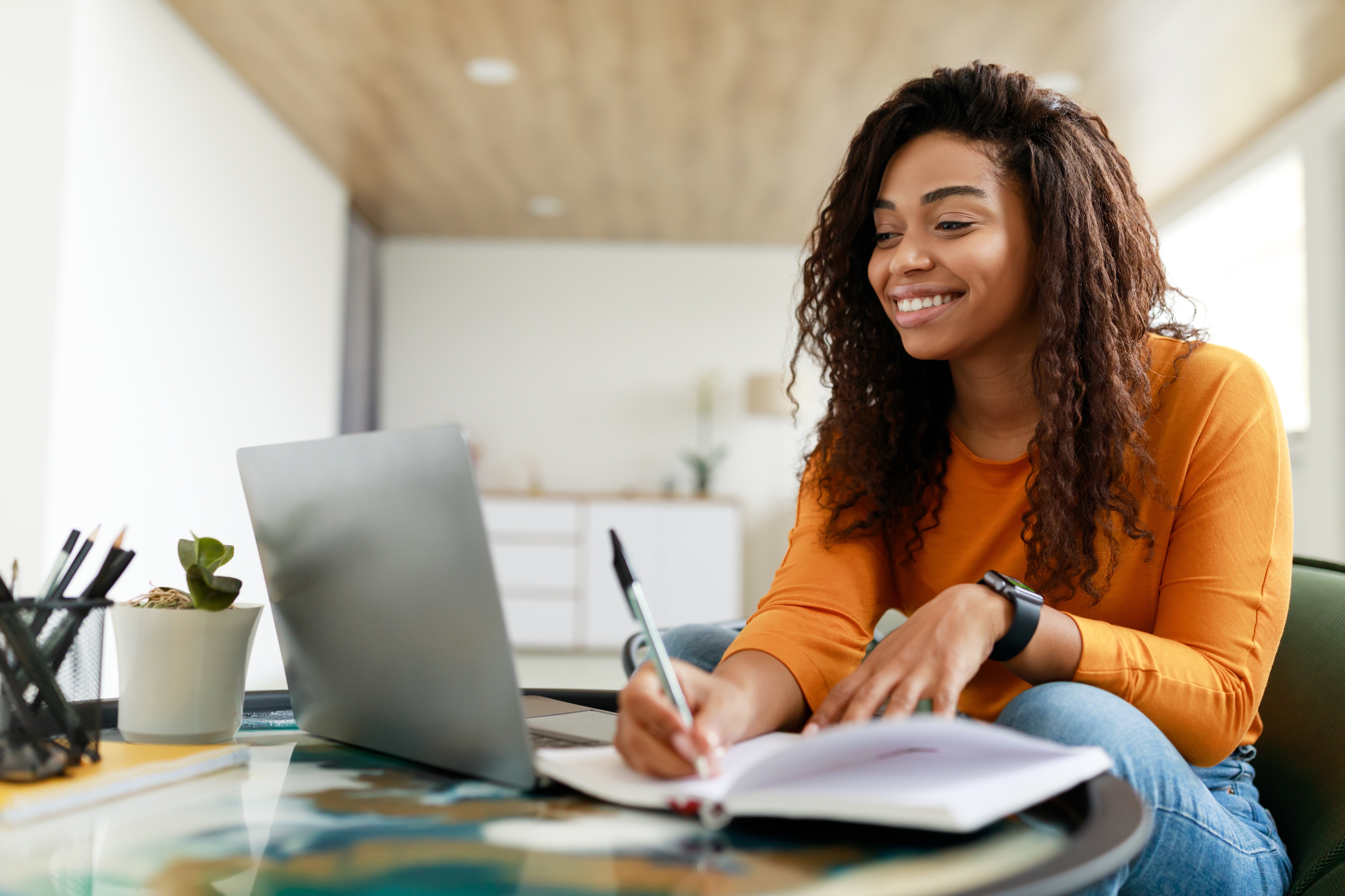 A woman with dark brown hair and brown skin is sat at a desk writing in a notepad and looking at a laptop screen. She is smiling as if she is on a video call. She appears to be working from home and is wearing casual clothes.