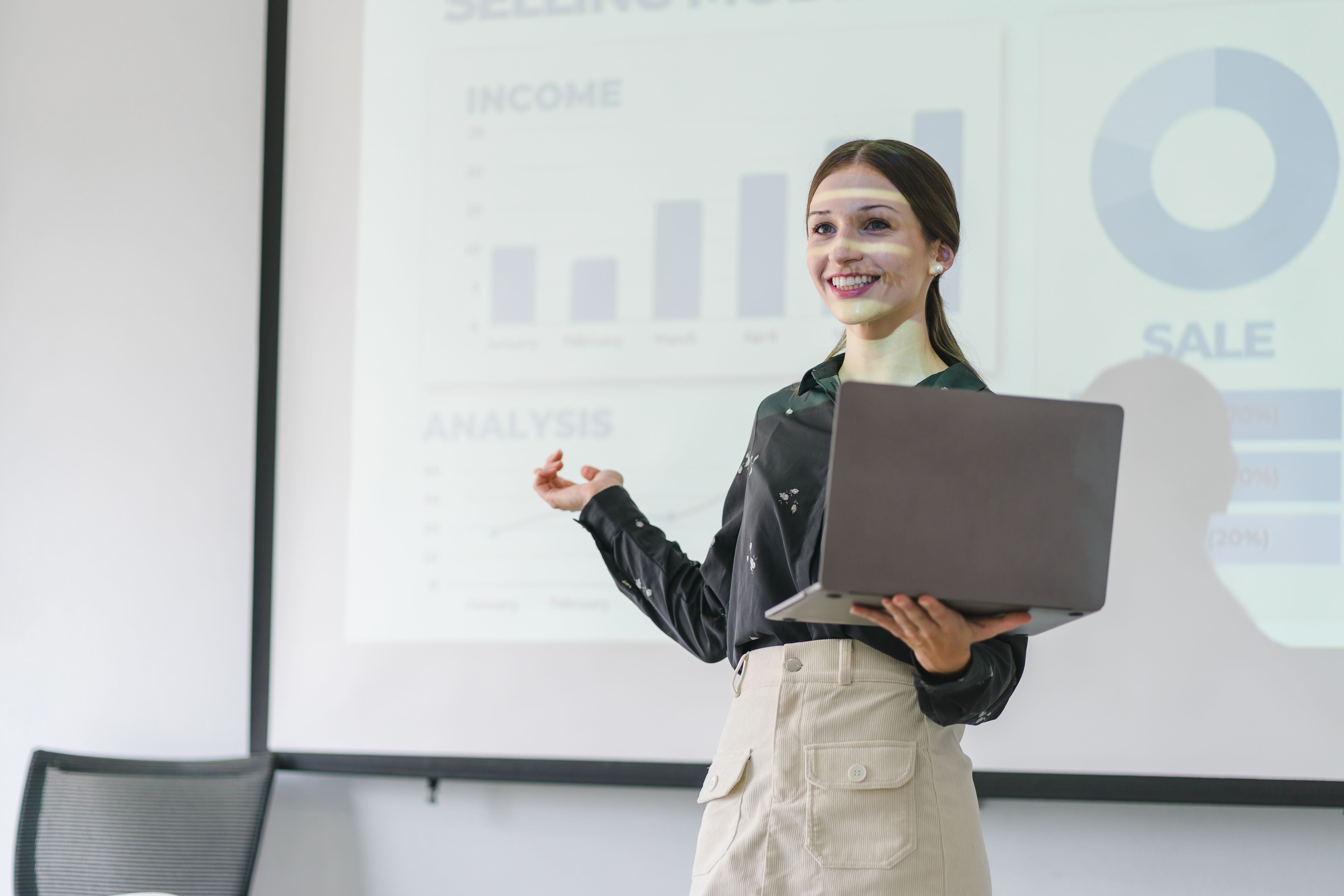 A professional woman is standing in front of a screen. She is holding a laptop and pointing to the screen behind her. She is smiling as if addressing and audience and presenting.