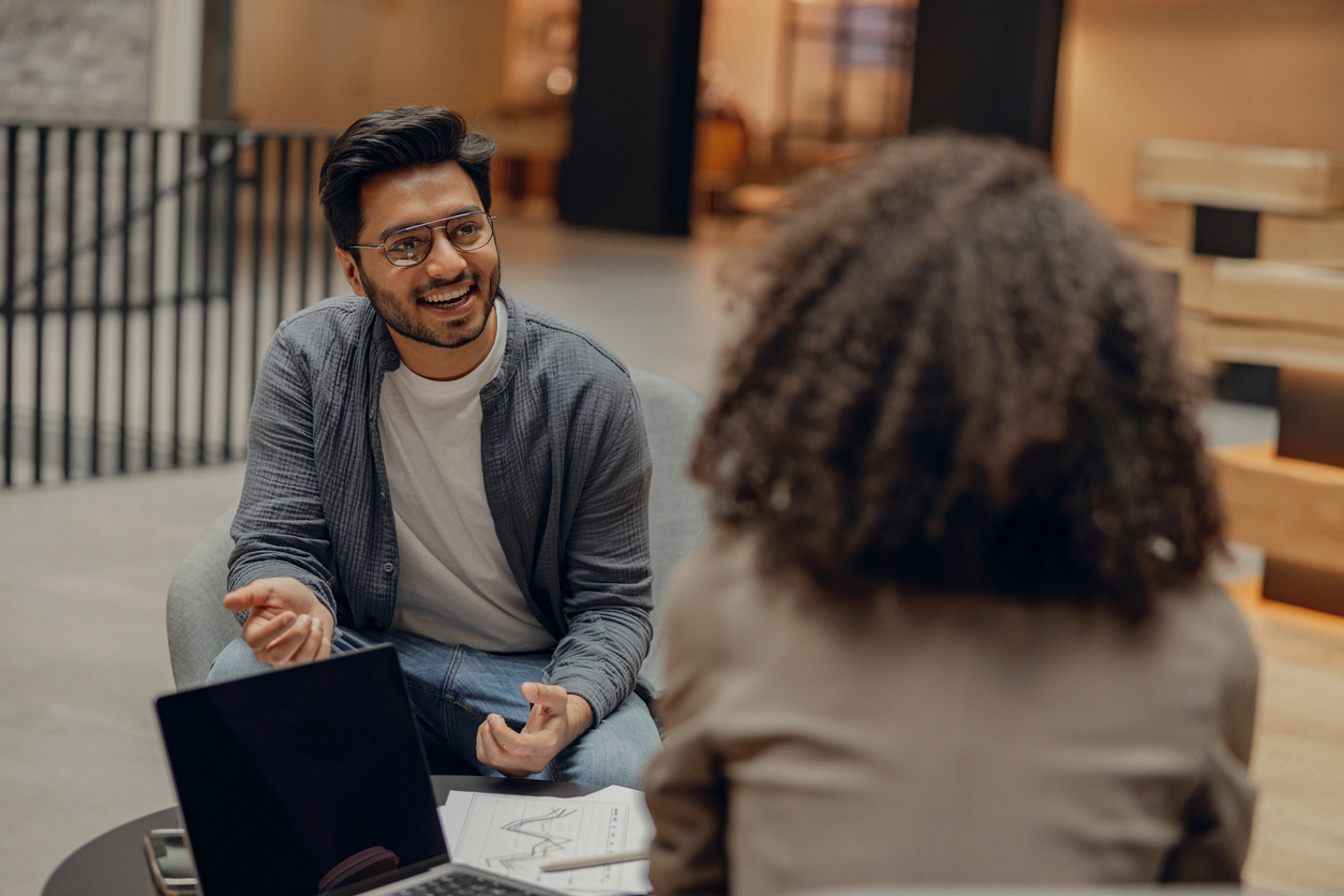 Two colleagues, a man and a woman, talking together in an office environment. There is a laptop and some paper notes on the table beside them and you can see the man is smiling and talking animatedly. 