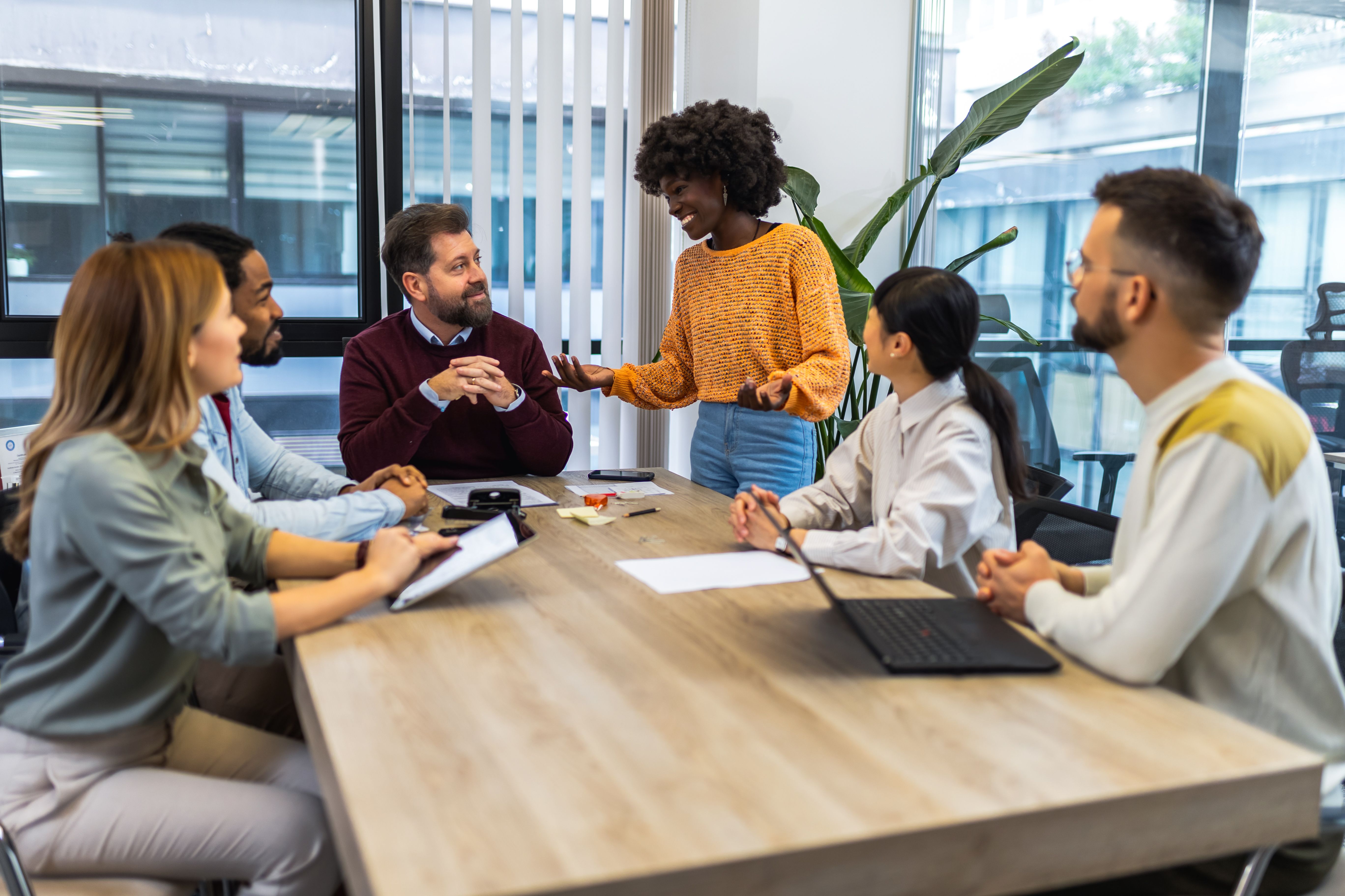 A group of colleagues gathered around a table in an office environment. A black woman wearing an orange jumper is standing and addressing her colleagues. She is smiling and they are smiling back. There are laptops and notepads on the table, suggesting they are in a meeting.