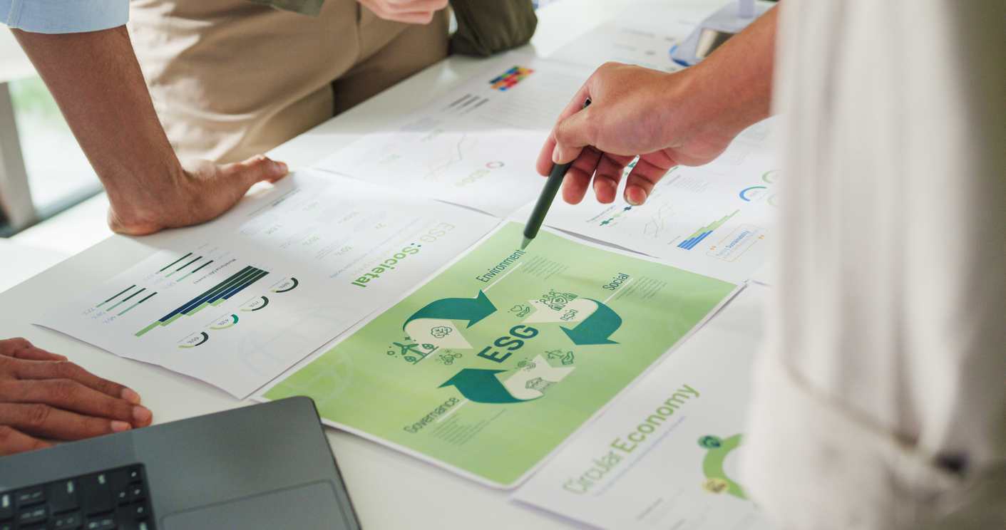 A number of printed slides are laid out on a table. The key topic of the slides is ESG. There are people stood around the table, pointing at the slides as if discussing them together. You can not see the people, only their hands pointing at the slides.