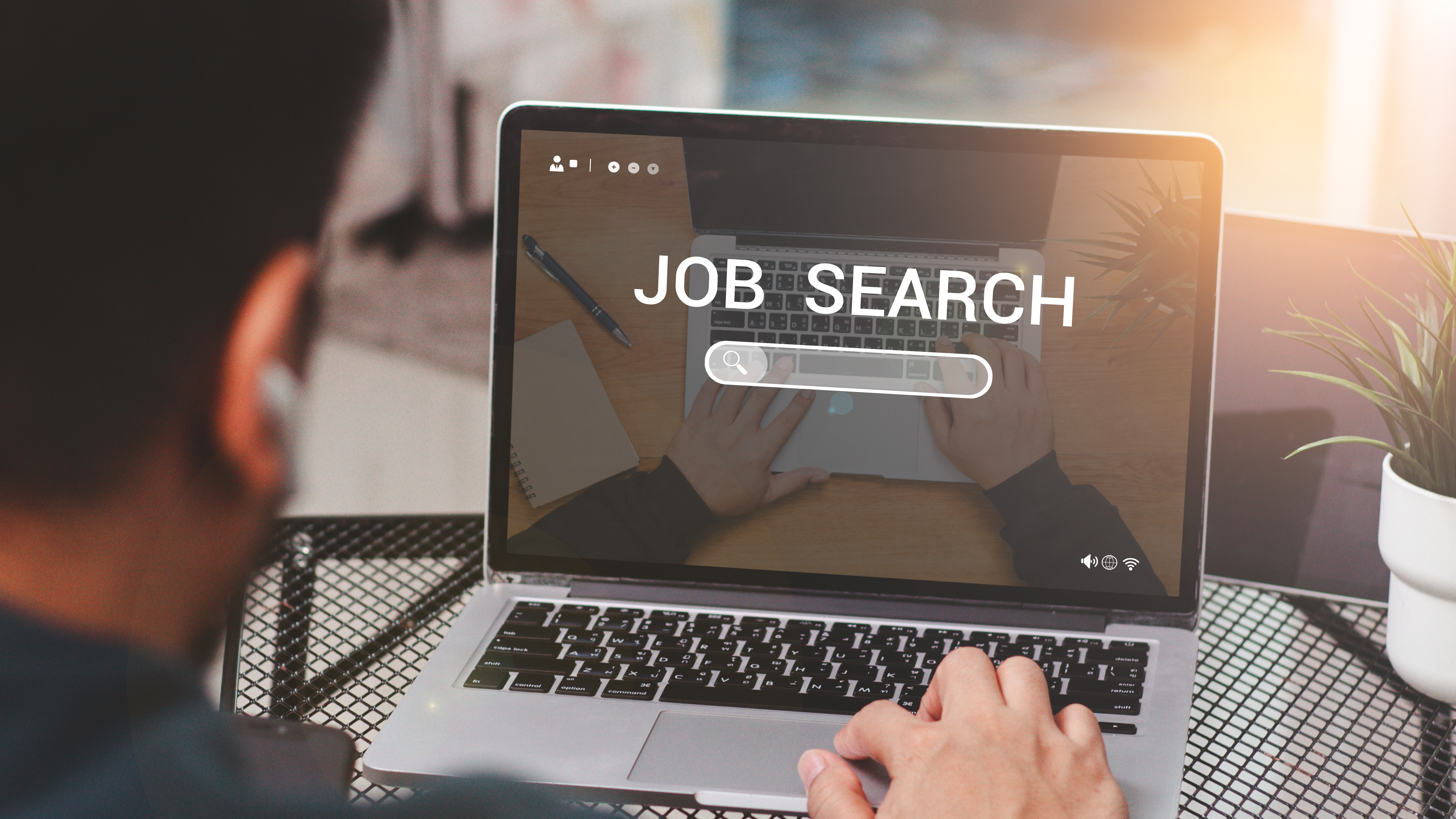 A man using a laptop. You can see the back of the man's head, his hand on the keyboard of the laptop and the laptop screen. The screen shows a job search website, indicating that the man is searching for a job.