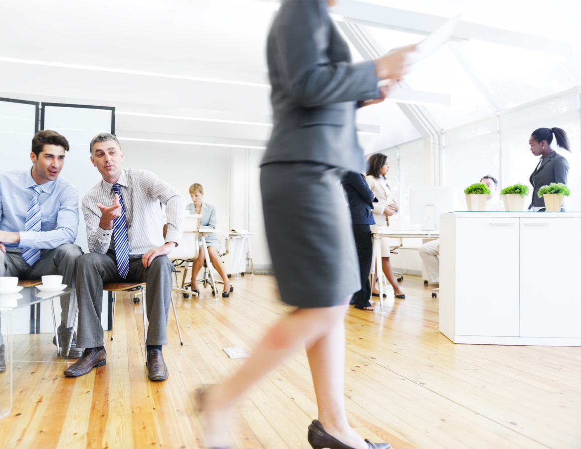 Colleagues in a workplace office environment. Two men are sat on chairs watching a female colleague walk past. The female colleague is wearing a smart skirt suit, and the men appear to be oggling her and making rude, suggestive remarks.