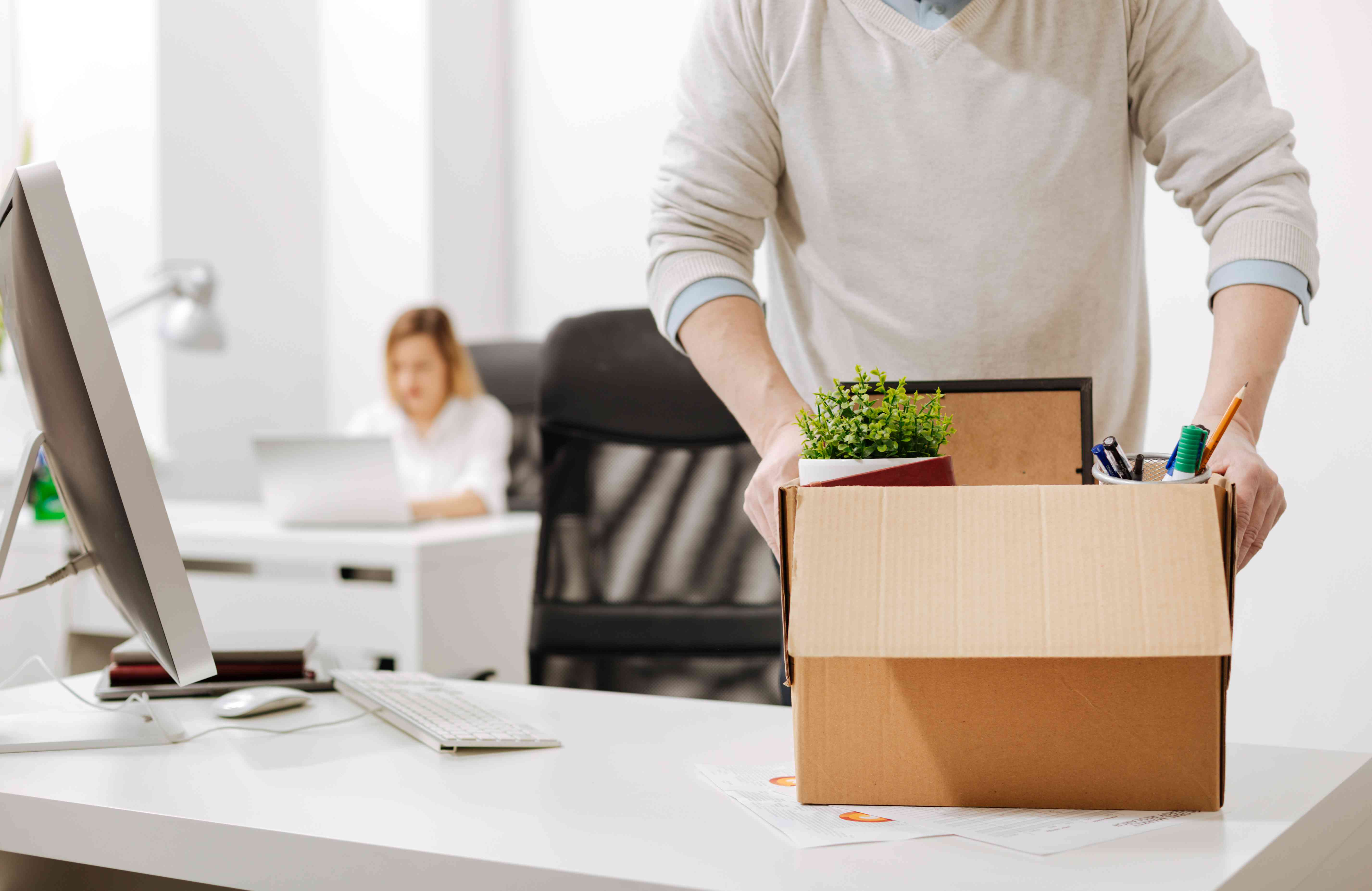 A person is stood at a desk in an office. There is a cardboard box in their hands that they have placed on the desk. The box has a plant, stationery and office supplies in. It looks as if the person is packing up their belongings to start a new job.