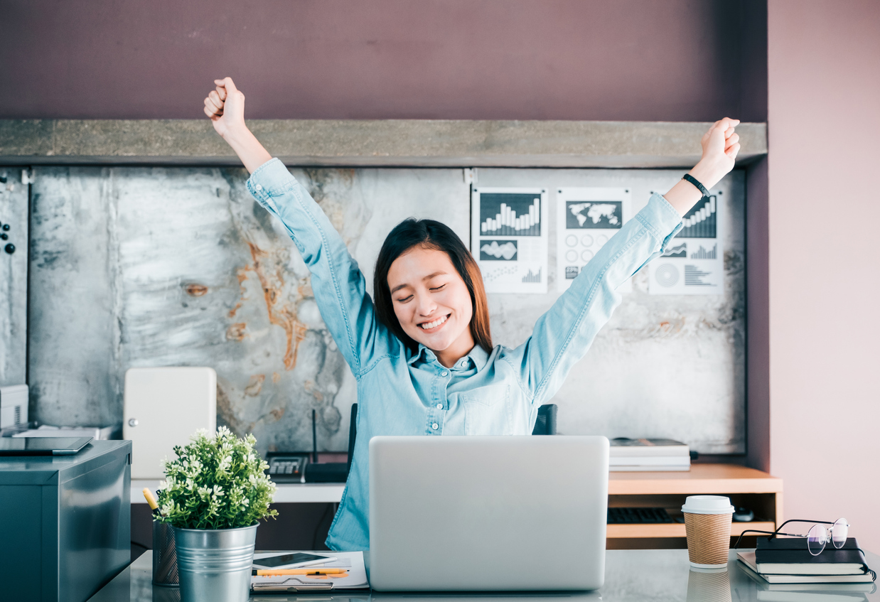 A woman is sat at a desk with a laptop in front of her. She is in an office environment, and there is a coffee cup, a plant, and a pile of books on the desk, too. She is smiling at her laptop screen and has her arms raised in the air. She looks happy and accomplished.