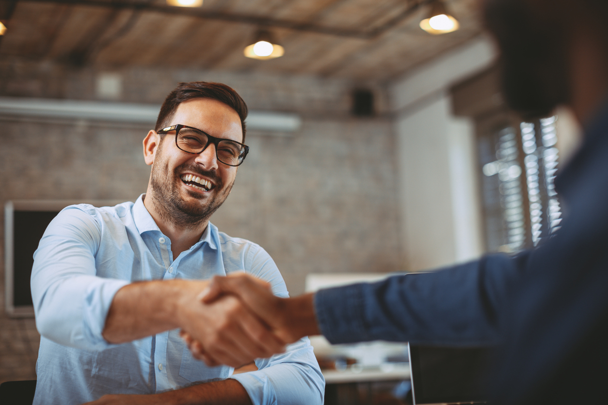 A man is smiling while shaking hands with another person who is out of shot. They are in a professional setting such as an office, and the man is wearing a blue shirt and glasses.