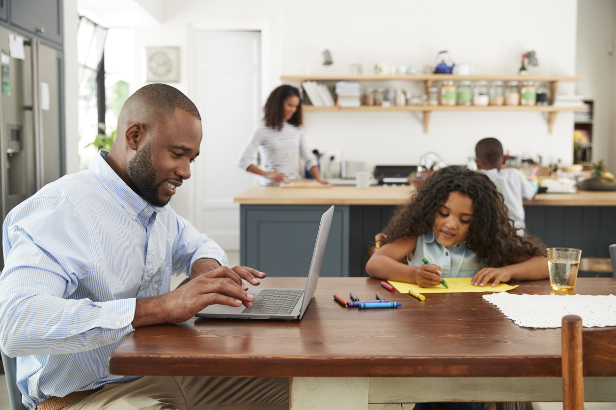 A family in their kitchen at home. A man is sat at the kitchen table working on a laptop, while a young girl is sat beside him colouring. In the background, a woman is stood at a counter preparing food while a young boy is also stood at the counter with her.