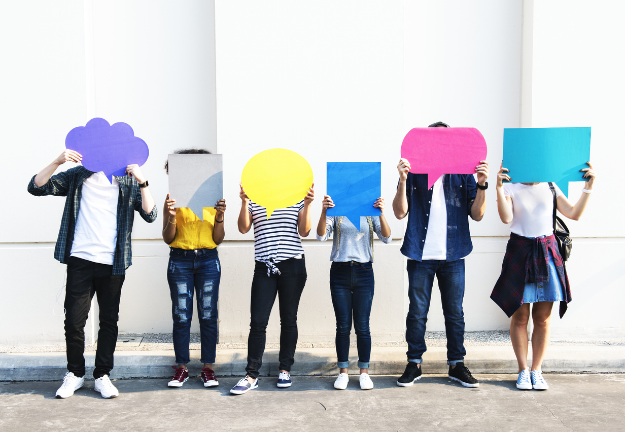 A photo of a diverse group of people, both men and women. They are standing against a wall outside, and are each holding a giant speech bubble in front of their faces.