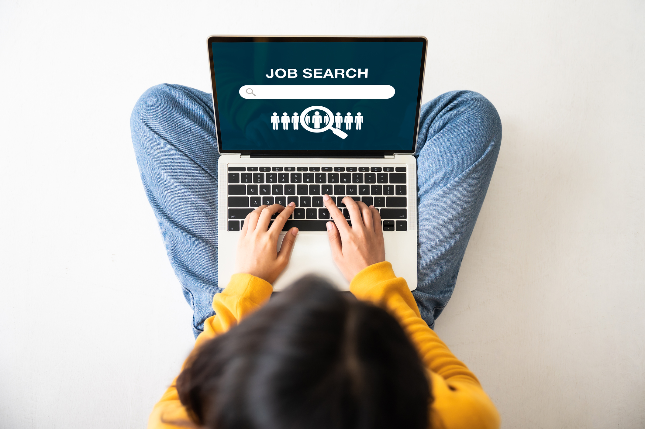 A woman is sat cross legged on the floor with a laptop on her lap. The laptop shows a job search bar, and she is typing as if she is looking for a new job.