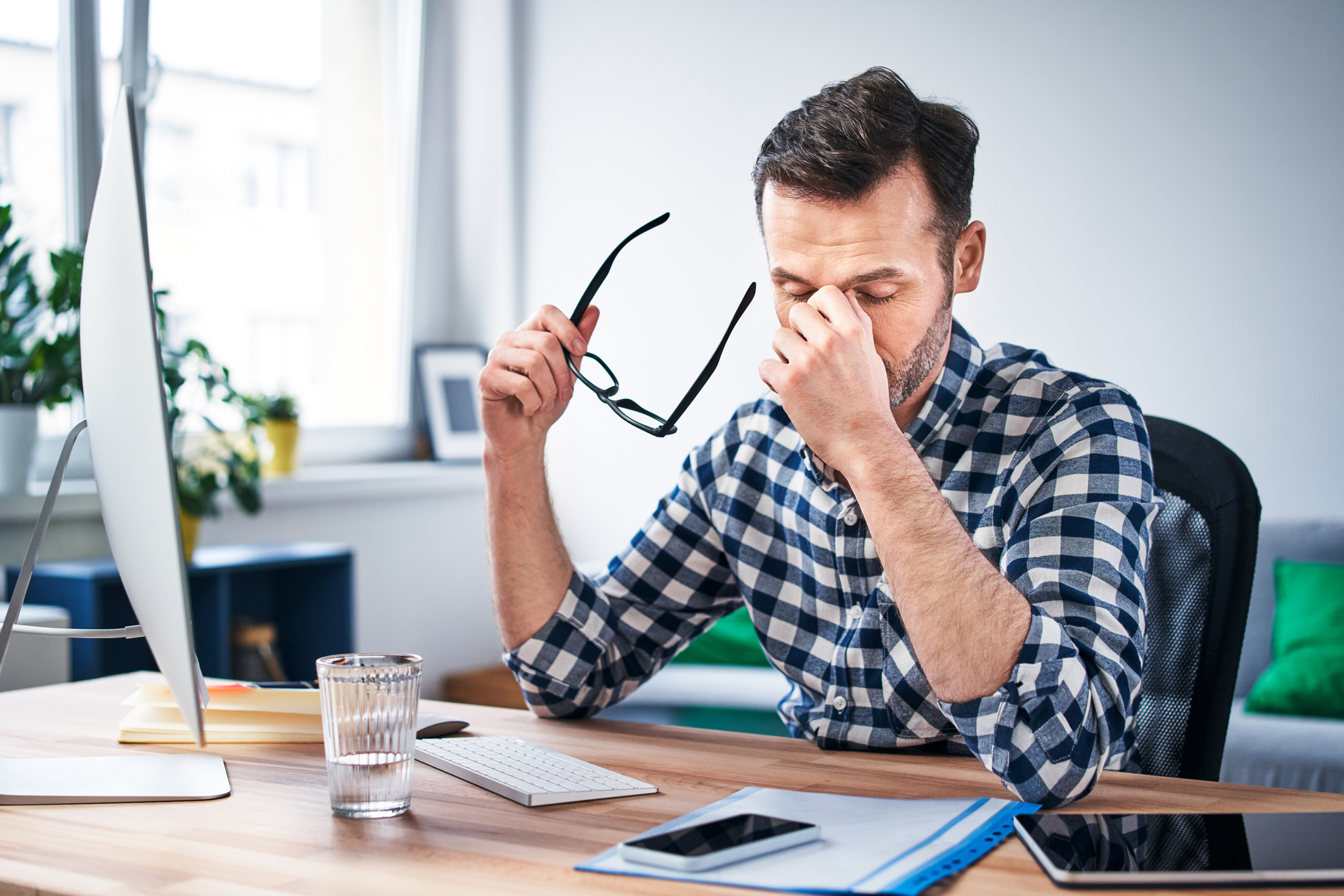 A photo of a man sat at a desk in front of a computer. He has his head in his hands and looks overwhelmed. 