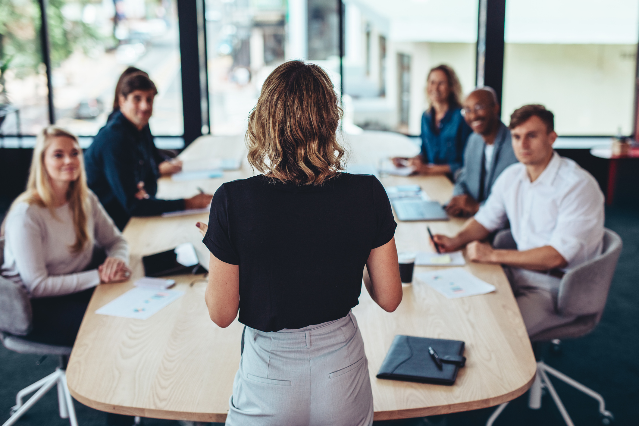 A photo of a woman standing at the head of a table in a boardroom. There are people sat around the table, looking at the woman as if listening to what she is saying.